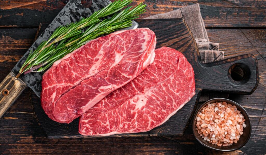 Două bucăți de umăr de vită, pe un tocător de lemn, lângă un bol cu sare grunjoasă roz și o legătură de rozmarin